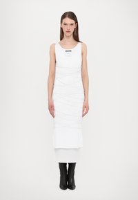 Rochie albă cu un design de top fără mâneci, având detalii din frânghie albă. Modelul poartă cizme negre până la genunchi. Eticheta de pe piept spune "Moschino."