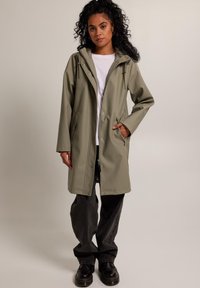 Pitkä, oliivinvihreä vedenpitävä takki, jossa on huppu, naru ja napit. Kerroksitettu valkoisen paidan päälle, yhdistetty tummiin farkkuihin ja mustiin saappaisiin.