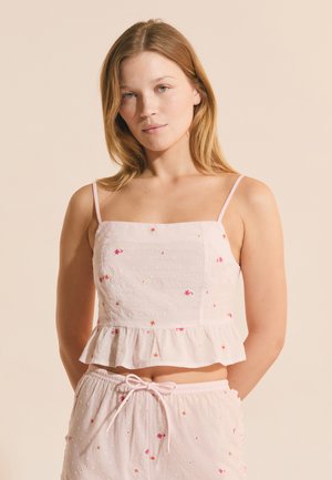 Młoda kobieta o jasnobrązowych włosach, ubrana w biały top bez rękawów oraz dopasowane krótkie spodenki z drobnymi różowymi kwiatowymi wzorami.