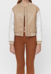 Chaqueta bomber beige de cuero sintético con mangas blancas, dobladillo y puños acanalados, cierre frontal con cremallera. El modelo lleva pantalones marrones con estampado.