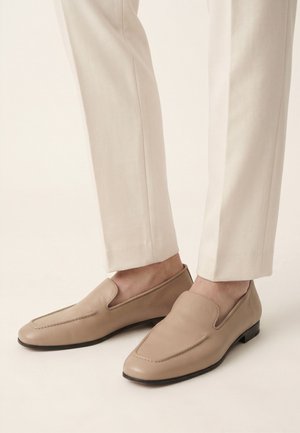 Beige læder slip-on loafers båret med lyse cremefarvede bukser, vist med nærbillede af fødder og underben på en neutral baggrund.