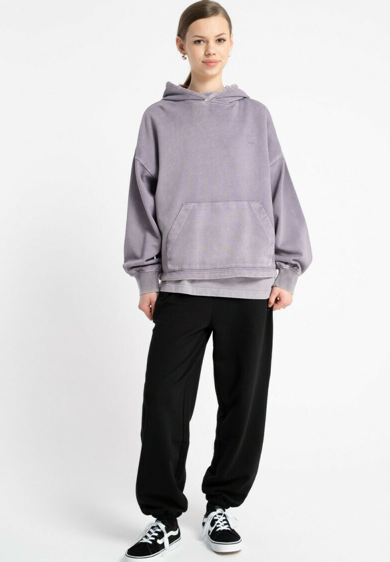Przesadzony fioletowy hoodie z przednią kieszenią, żebrowanymi mankietami i dołem, w połączeniu z luźnymi czarnymi spodniami i czarnymi trampkami.