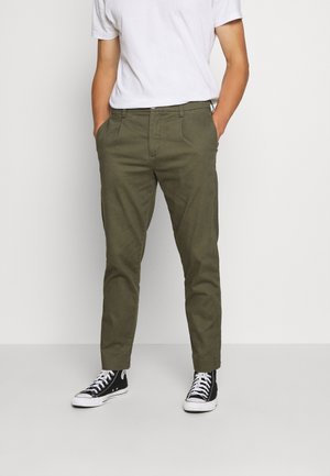Personne portant un pantalon vert olive, un t-shirt blanc et des baskets montantes noires, debout les mains dans les poches devant un fond uni.