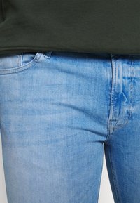 Vaqueros de mezclilla de lavado claro con una textura suave, que presentan un diseño clásico de cinco bolsillos y detalles sutiles de costura. Combinados con una camiseta de color verde oscuro.