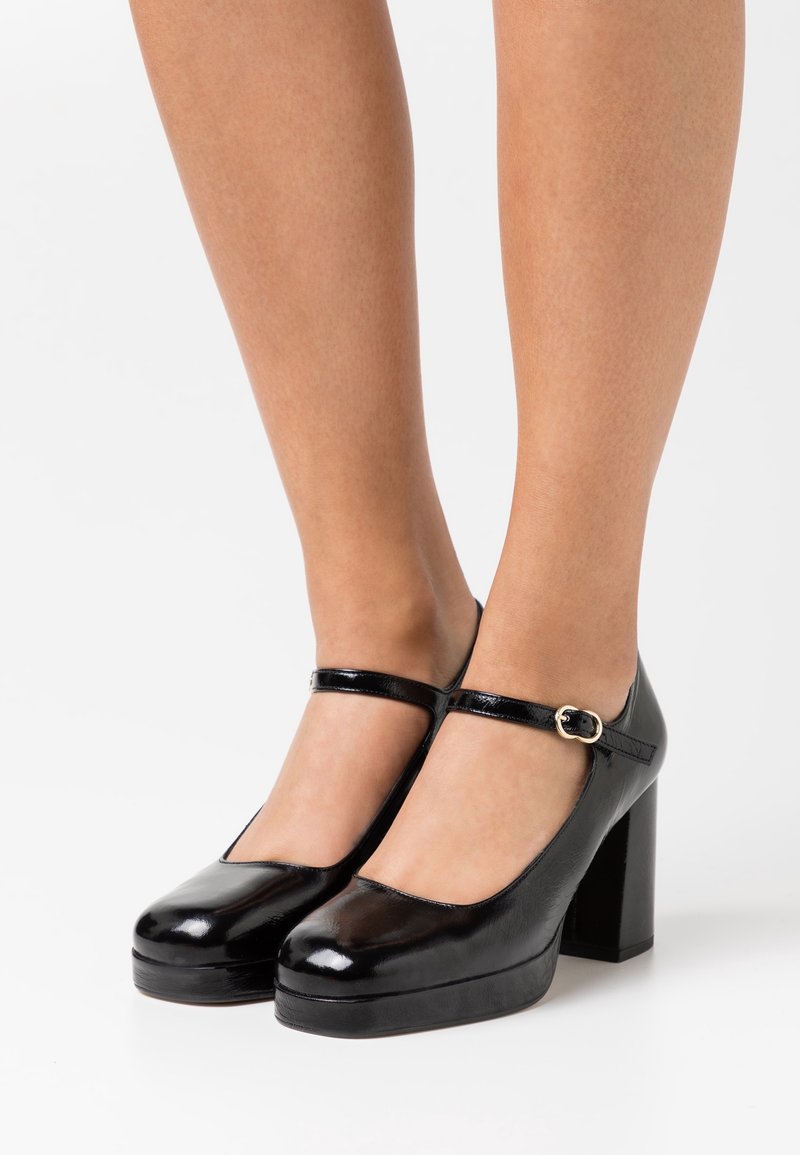 Des jambes portant des escarpins Mary Jane en cuir verni noir avec des lanières à la cheville et des talons blocs sur fond blanc.