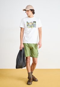 Weißes Baumwoll-T-Shirt mit Grafikdruck, grüne Cargo-Shorts, graue Pufferjacke, braune Sandalen und beiger Cap auf einem gelben Boden.