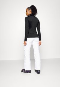 Schwarzes Langarmoberteil mit Stehkragen, kombiniert mit weißen Hosen. Das Outfit zeichnet sich durch klare Linien und eine taillierte Silhouette ohne sichtbare Muster aus.
