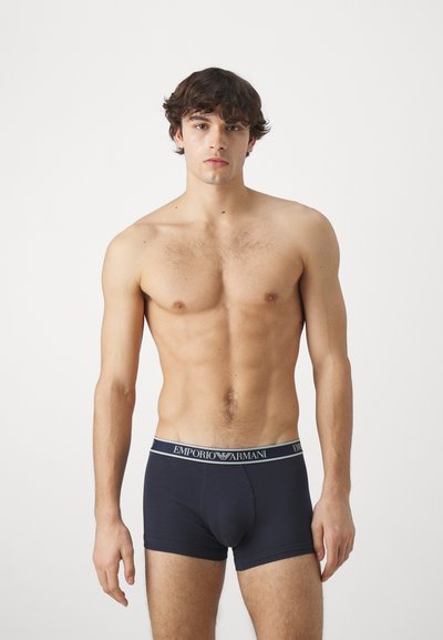 Námornícky modré bavlnené boxerky s jemným pásom, na ktorom sa nachádza logo "Emporio Armani" v bielej a striebornej farbe. Model stojí s rukami pozdĺž tela.