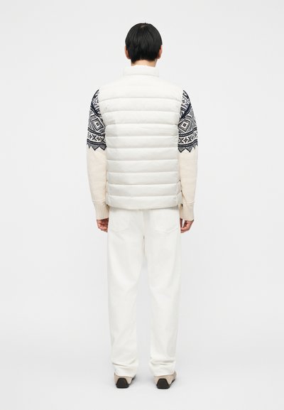 Λευκό γιλέκο puffer με σχέδια σε темноμπλε μανίκια, φορεμένο πάνω από μπεζ πουλόβερ και κρεμ παντελόνι. Λείος υφασμάτινος, κεντημένος σχεδιασμός και άνετη εφαρμογή.