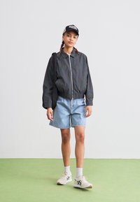 Svart bomberjakke med glidelås foran, lyse denimshorts og hvite joggesko. Modellen har også på seg en svart caps med logo.