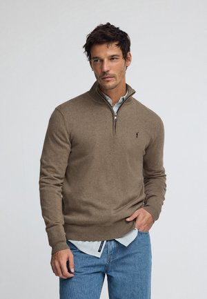 Brown knit pullover with a ribbed collar and cuffs, featuring a front zipper and embroidered logo. Worn over a light blue collared shirt.