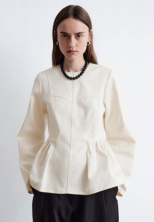 Cream peplum blouse with long sleeves, featuring a structured design, round neckline, and subtle stitching details at the bodice. Black bead necklace.