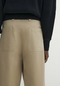 Black ribbed sweater tucked into high-waisted beige pants with two large square back pockets, shown from waist to mid-thigh.