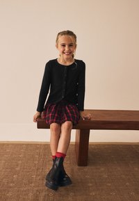 Haut noir à manches longues avec boutons, jupe écossaise rouge et bottines noires à semelles épaisses. Enfant assis sur un banc en bois.