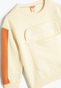 Sudadera color crema con textura acanalada, que presenta un acento naranja en la manga y un gran bolsillo frontal. Tejido suave y escote redondo.