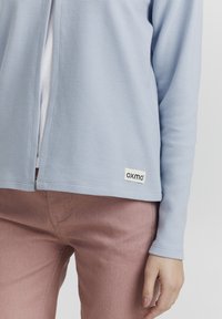 Svijetloplavi rebrasti zip-up jakna izrađena od teksturirane tkanine, s dugim rukavima i bijelom etiketom s natpisom "oxmo" u crnoj boji. Uparena s ružičastim trapericama.