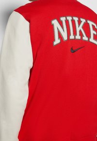 Chaqueta estilo universitario en rojo y blanco con la palabra "NIKE" bordada, textura suave y un pequeño detalle con logo negro.