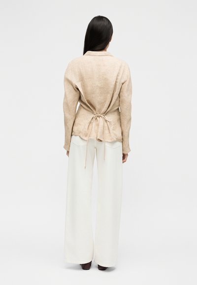Γυναίκα που στέκεται με την πλάτη στραμμένη προς τον θεατή, φοράει μια μπεζ μπλούζα με μακριά μανίκια δεμένη στη μέση και φαρδιά λευκή παντελόνα, μπροστά σε απλό φόντο.