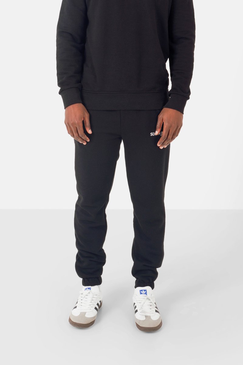 Osoba ubrana w czarne dresy i czarną bluzę, stojąca, w białych i czarnych sneakersach Adidas na białym tle.