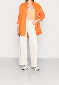 En överdimensionerad orange skjorta över en ljus persikofärgad t-shirt med grafik. Vita vida byxor och ljusa sneakers fulländar looken.