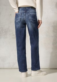 Jeans de mezclilla azul oscuro con corte recto, que presentan bolsillos traseros y un desvanecimiento sutil. Combinados con zapatillas blancas.