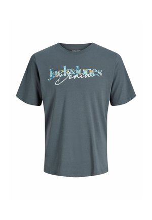 Lyshåret dreng står og smiler iført en grå t-shirt med grafik, vide blå jeans og beige sko mod en ensartet lys baggrund.