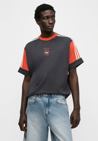 Γκρι υπερμεγέθη κοντομάνικη μπλούζα με κόκκινες λεπτομέρειες, διακοσμημένη με λευκές ρίγες στους ώμους και κεντημένο λογότυπο. Συνδυάζεται με ανοιχτόχρωμα τζιν παντελόνια.