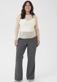 Hellbeige Bluse mit langen Ärmeln und glitzernden Punkten, über einem eng anliegenden cremefarbenen Tanktop getragen, kombiniert mit grauen weiten Hosen. Silberne Akzente an den Schuhen.