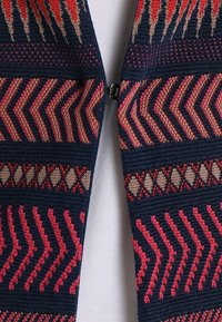 Strickjacke mit marineblauem Grund, die geometrische Muster in Rot und Beige aufweist. Offenes Frontdesign mit einem metallischen Reißverschlussdetail.