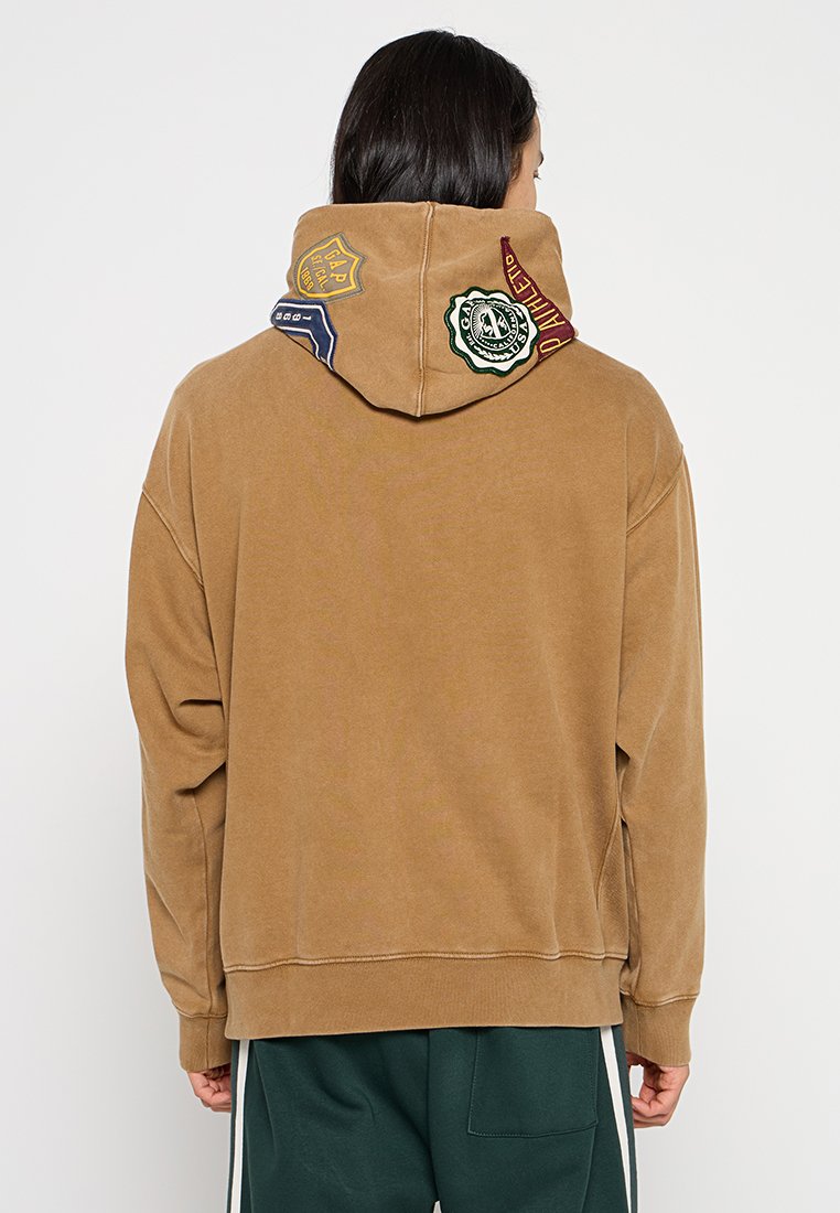 Brauner, übergroßer Hoodie mit strukturierter Oberfläche, der bunte Patches an der Kapuze und einen gerippten Saum aufweist. Weicher Stoff, lässige Passform.