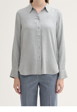 Person iført en lysegrå langærmet skjorte med knapper og mellemgrå, skræddersyede bukser, stående foran en ensfarvet baggrund.