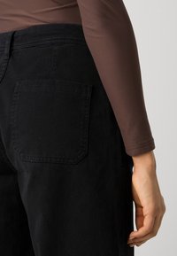 Črne jeans hlače z zadnjim žepom in visokim pasom, v kombinaciji z dolgo rjavo majico. Tkanina ima gladko teksturo.