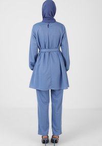 Blaue Tunika mit tailliertem Gürtel und langen Ärmeln, kombiniert mit passenden Hosen. Glattes Material, lockere Passform und ein Knopfdetail am Hals.