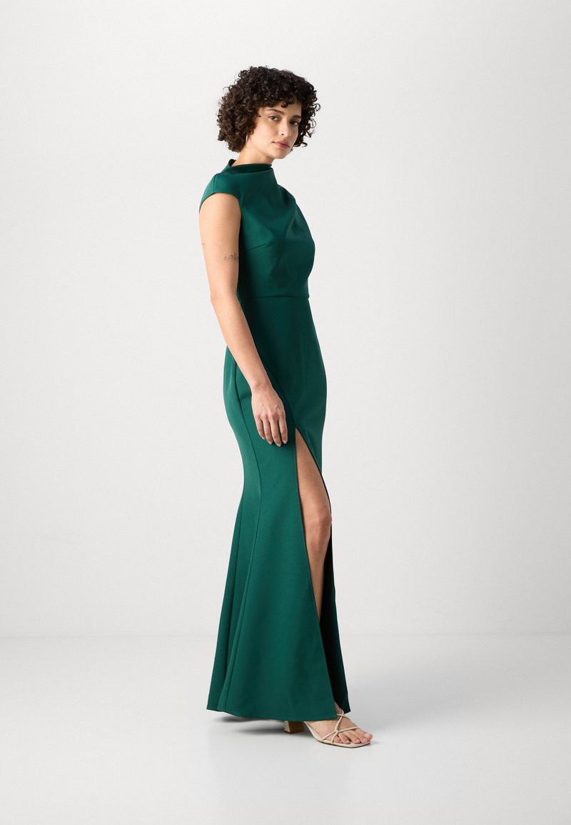 Zielona wieczorowa suknia z gładkiego materiału, z dopasowanym gorsetem, rękawami typu cap i rozcięciem sięgającym do uda. W połączeniu z sandałami na otwartym palcu.