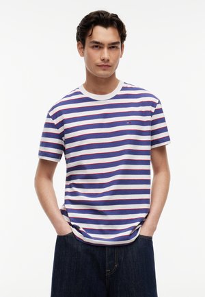 Młody mężczyzna o ciemnych włosach, ubrany w niebiesko-biało-czerwoną koszulkę w paski z krótkim rękawem i ciemne spodnie, ręce w kieszeniach, neutralne tło.