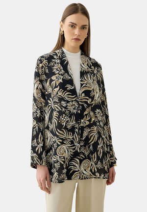 Frau trägt einen schwarzen Blazer mit goldenen und beigen Blumenmustern über einem weißen Top und beigen Hosen vor einem schlichten Hintergrund.