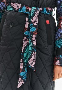 Čierny prešívaný kabát s opaskom z látky s kvetinovým vzorom. Má kontrastný gombík a živé motívy listov v ružovej a modrej farbe.