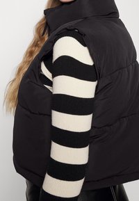 Czarna pikowana kamizelka z wysokim kołnierzem, o gładkiej fakturze, noszona na czarno-białym swetrze w paski.