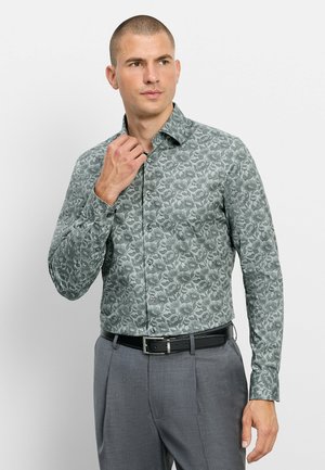 Man past de kraag aan van een groen bloemetjespatroon overhemd met lange mouwen, draagt een grijze broek en een zwarte riem, kijkt opzij.