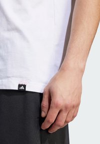 T-shirt bianco in cotone con un'etichetta nera del logo Adidas sull'orlo, caratterizzato da dettagli di cucitura rosa. Indossato con pantaloni corti scuri.