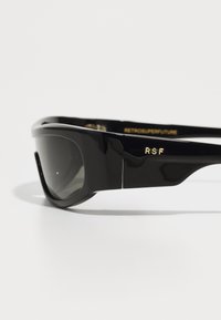 Black sunglasses with a sleek design, featuring dark lenses, a glossy finish, and gold detailing on the temple that reads "RSF" and "RETROSUPERFUTURE."