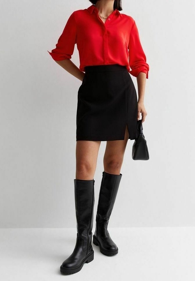 Chemise rouge à boutons avec manches retroussées, mini-jupe noire fendue sur le côté, et bottes en cuir noires jusqu'aux genoux. Petit sac à main noir inclus.