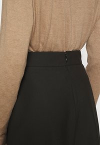Haut marron à manches longues rentré dans une jupe noire taille haute avec une fermeture éclair latérale, vue de dos au niveau de la taille.
