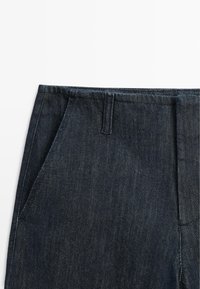 Dunkelblaue Jeanshose mit sichtbarer Gürtelschlaufe und seitlicher Tasche auf weißem Hintergrund.