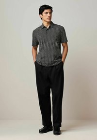 Camicetta nera a maniche corte con motivi geometrici bianchi, abbinata a pantaloni neri larghi e scarpe nere, presentata su uno sfondo neutro.