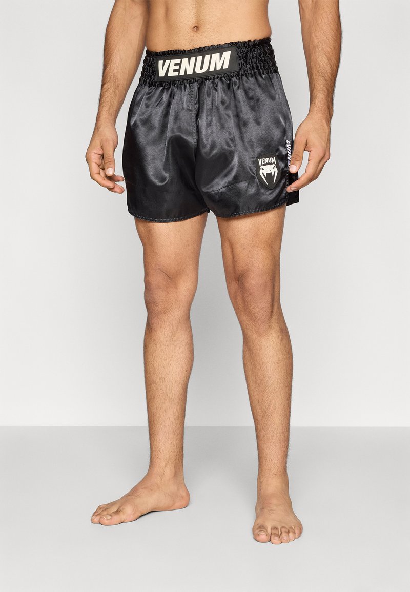 Calções pretos em cetim com cós elástico apresentando um logótipo "Venum" bege e um gráfico de logótipo no lado esquerdo. Bainha dobrada e design leve.