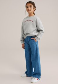 Siv dolg memento z rdečim napisom, v kombinaciji z ohlapnimi modrimi kavbojkami. Bele športne copate z rdečimi in modrimi detajli. Preprost, sproščen outfit.