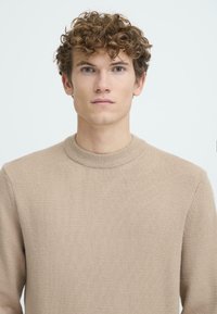Beige Pullover mit Rundhalsausschnitt aus weichem Strickstoff. Verfügt über einen gerippten Kragen und Ärmelbündchen sowie eine gleichmäßige Textur.