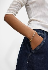 Złota i srebrna bransoletka o smukłym, płaskim designie z rzędem przezroczystych kamieni. Noszona na ręce spoczywającej w kieszeni ciemnoniebieskich dżinsów.