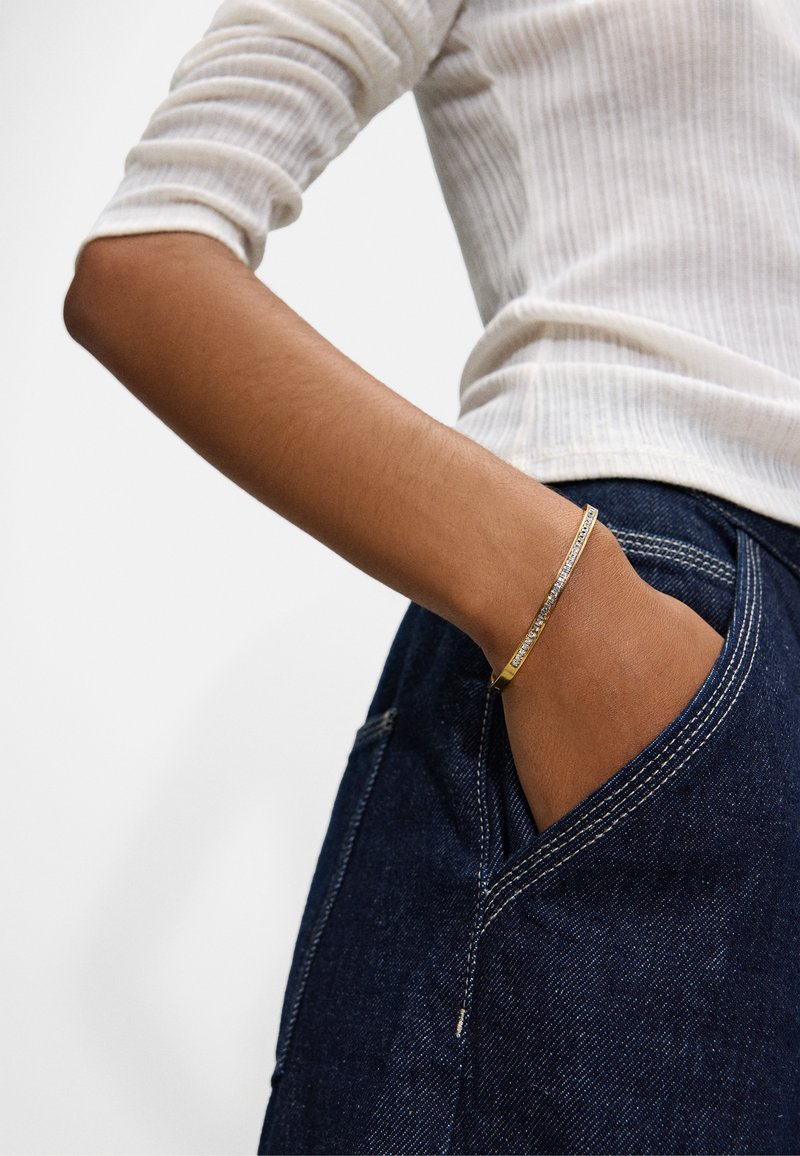 Złota i srebrna bransoletka o smukłym, płaskim designie z rzędem przezroczystych kamieni. Noszona na ręce spoczywającej w kieszeni ciemnoniebieskich dżinsów.